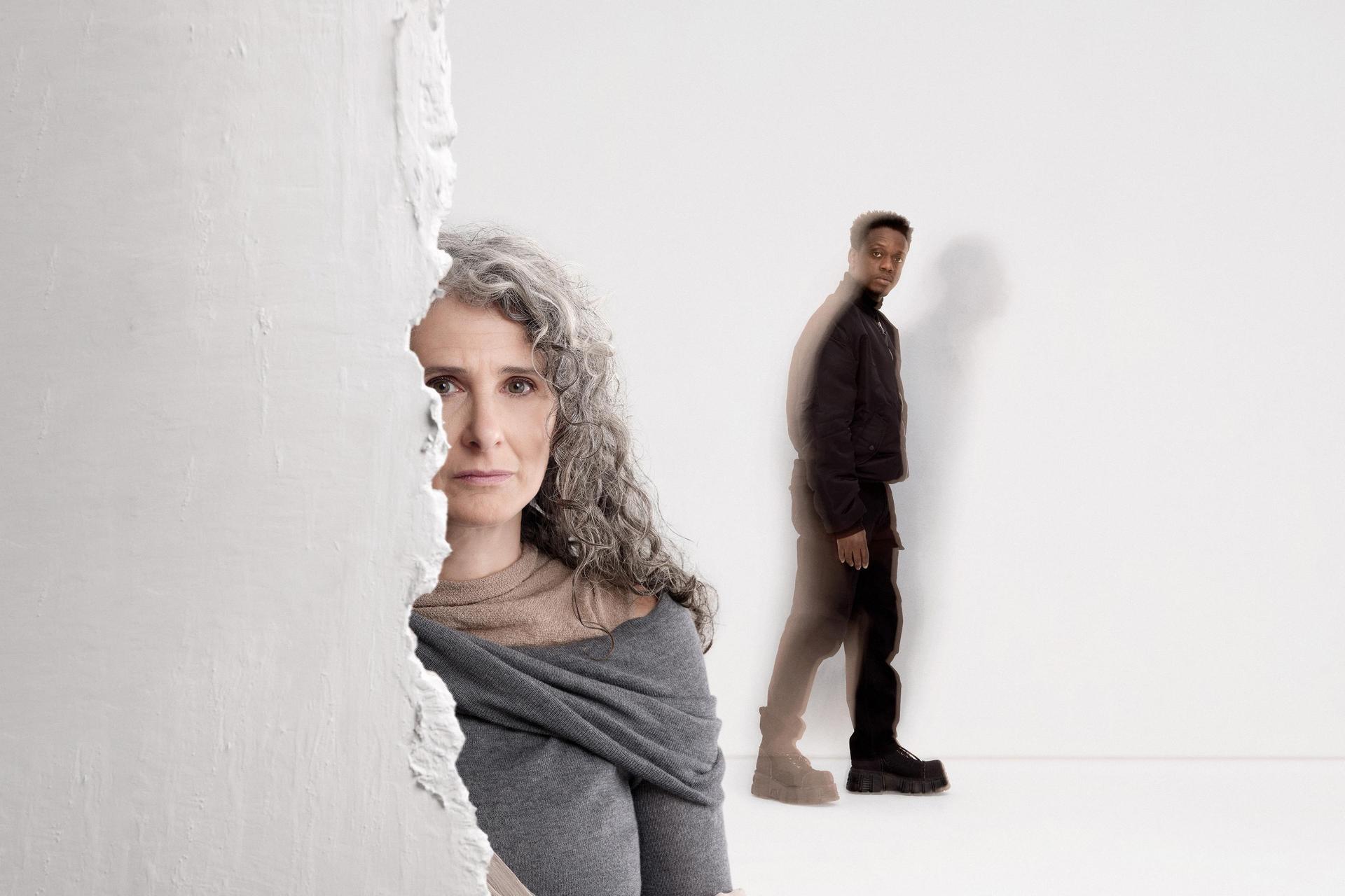 Vrouw in het grijs gekleed staat half achter muur met jongen in het zwart gekleed op de achtergrond.